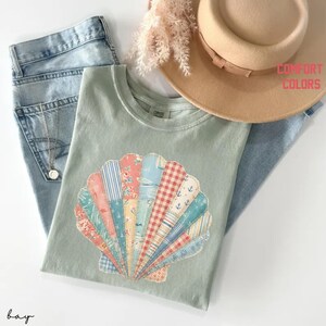 May include: A light green t-shirt featuring a seashell design made of various patterned fabrics. The shirt is paired with blue jeans and a tan hat. The words "COMFORT COLORS" are printed on the upper right.