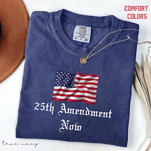 May include: A true navy blue t-shirt with the text "25th Amendment Now" and an American flag graphic. The shirt is a Comfort Colors brand and has a crew neck. A gold necklace is draped across the shirt.