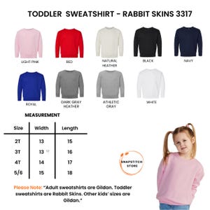 K&ouml;nnte beinhalten: Eine Auswahl an Sweatshirts f&uuml;r Kleinkinder in verschiedenen Farben, darunter Hellrosa, Rot und Marineblau. Das Bild zeigt eine Gr&ouml;&szlig;entabelle mit Ma&szlig;en in Zentimetern und den Text "TODDLER SWEATSHIRT - RABBIT SKINS 3317". Ein junges M&auml;dchen tr&auml;gt ein hellrosa Sweatshirt.
