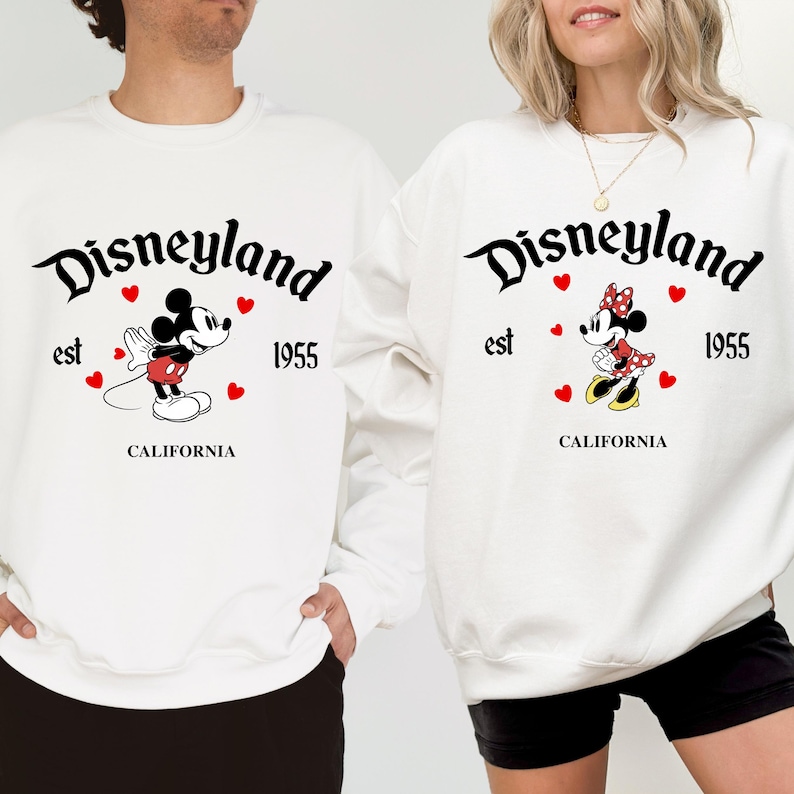 Pode incluir: Camisolas brancas com o logotipo da Disneyland, gr&aacute;ficos do Mickey Mouse e da Minnie Mouse. As camisolas t&ecirc;m o texto "Disneyland est 1955 CALIFORNIA" em escrita preta. Cora&ccedil;&otilde;es vermelhos est&atilde;o espalhados pelos personagens.