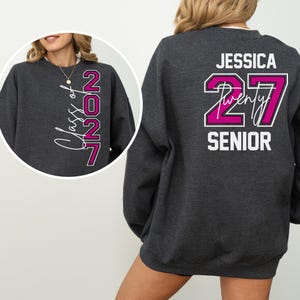 May include: A dark gray sweatshirt with white and magenta text. The front features "Class of 2027" in a script font. The back displays "Jessica" above "27 Twenty Senior" in a bold, outlined font. A casual, comfortable garment.