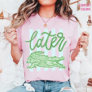 May include: Pink t-shirt with the word "later" in green script, over a green alligator illustration. The shirt has light blue and pink vertical stripes in the background. The words "Comfort Colors" are in the upper right corner.