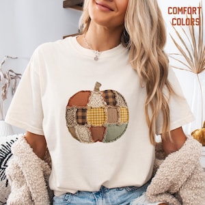 May include: A cream-colored short-sleeve t-shirt featuring a central graphic of a pumpkin composed of various fabric patches in shades of brown, tan, yellow, and sage green, including leopard print, gingham, and plaid patterns, with the text COMFORT COLORS.