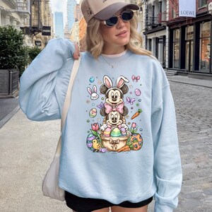 K&ouml;nnte beinhalten: Hellblaues Sweatshirt mit einem Ostermotiv. Das Design zeigt Mickey und Minnie Mouse-Charaktere mit Hasenohren, Ostereiern, Schmetterlingen und Karotten. Das Sweatshirt hat lange &Auml;rmel und einen Rundhalsausschnitt.