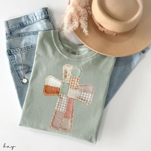 May include: A pale green t-shirt featuring a patchwork cross design in various colours and patterns. The shirt is laid with light blue jeans and a tan hat. The word "bay" is written in the bottom left corner.