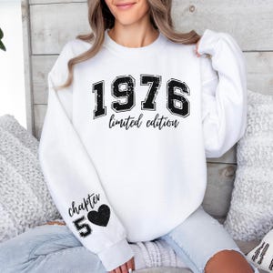 May include: A white crewneck sweatshirt featuring the black distressed text 1976 limited edition on the chest. The right sleeve displays the black text chapter 50 above a solid black heart icon. The garment has long sleeves and a ribbed neckline.