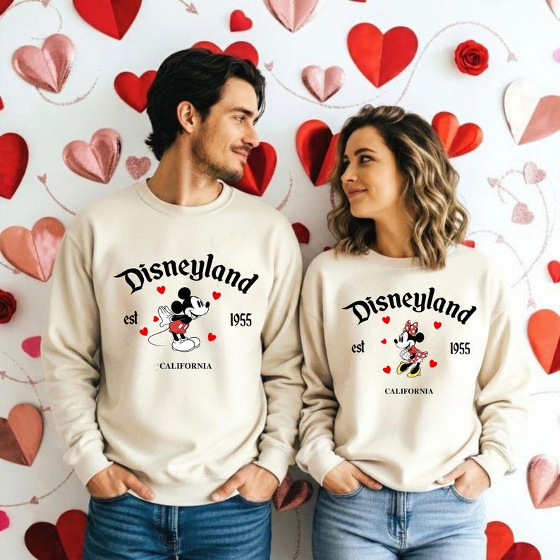 Pode incluir: Duas camisolas bege com o log&oacute;tipo da Disneyland, gr&aacute;ficos do Mickey Mouse e da Minnie Mouse, e o texto "est 1955 CALIFORNIA". O fundo apresenta decora&ccedil;&otilde;es de cora&ccedil;&otilde;es vermelhos e rosa.