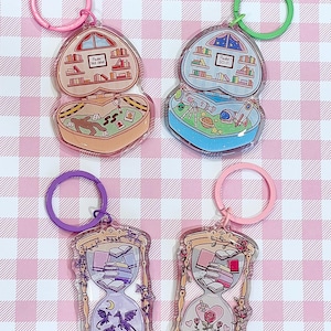 Może przedstawiać: Cztery akrylowe breloczki do kluczy. Dwa są w kształcie serca, przedstawiające bibliotekę i miejsce zbrodni. Pozostałe dwa mają kształt klepsydry, z stosami książek i wzorami kwiatowymi lub smokami. Breloki są różowe, zielone i fioletowe.