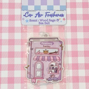 May include: A car air freshener in a clear plastic package. The air freshener is shaped like a purple fantasy bookstore with the text "Fantasy Bookstore". The top of the package has the text "Car Air Freshener" and "Scent: Wood Sage & Sea Salt".
