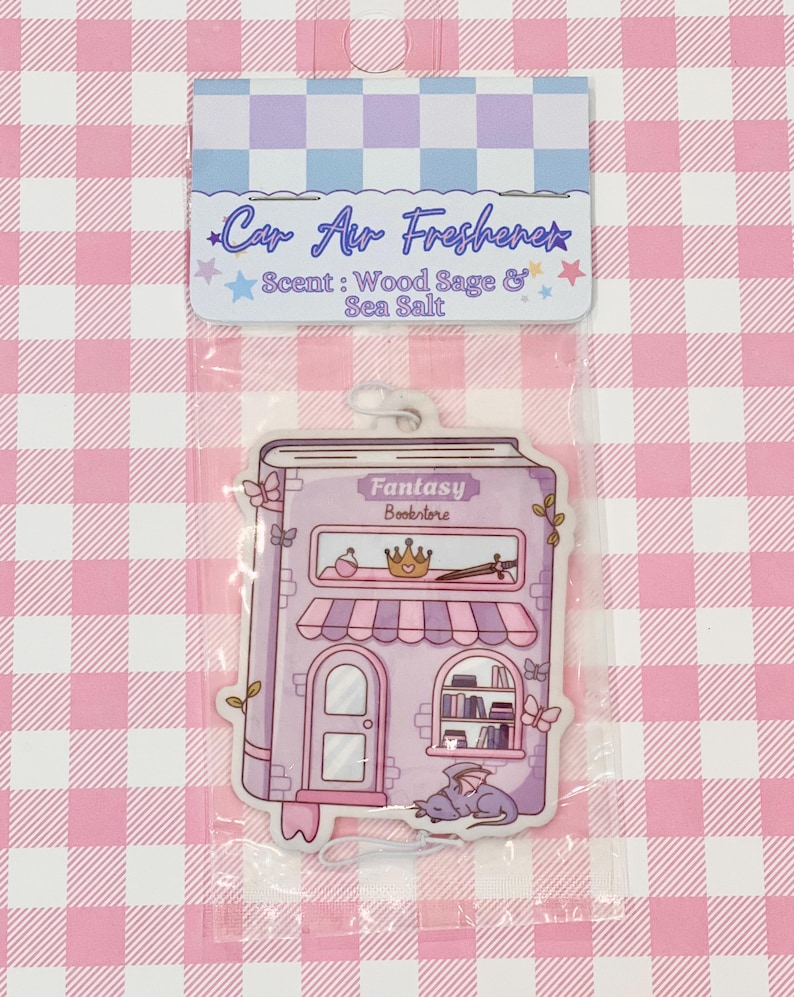 May include: A car air freshener in a clear plastic package. The air freshener is shaped like a purple fantasy bookstore with the text "Fantasy Bookstore". The top of the package has the text "Car Air Freshener" and "Scent: Wood Sage & Sea Salt".