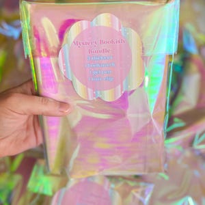 以下が含まれることがあります： 「Mystery Bookish Bundle」が入った透明な虹色のパッケージ。バンドルには、ステッカー3枚、ブックマーク1枚、ジェルペン1本、ヘアクリップ1個が含まれています。ラベルはパステルカラーで、「Mystery Bookish Bundle」と書かれています。
