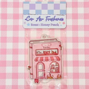 May include: A pink car air freshener shaped like a book, with the text "Spicy Bookstore." The top of the packaging reads "Car Air Freshener" and "Scent: Honey Peach."
