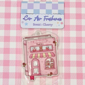 May include: Pink car air freshener in a clear plastic package. The air freshener is shaped like a pink bookstore with the words "Romance Bookstore" on the front. The packaging reads "Car Air Freshener" and "Scent: Cherry".