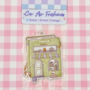 May include: Car air freshener in a clear plastic package. The air freshener is shaped like a green bookstore with the text "Thriller Bookstore" and "Scent: Sweet Orange". The background is a pink and white checkered pattern.