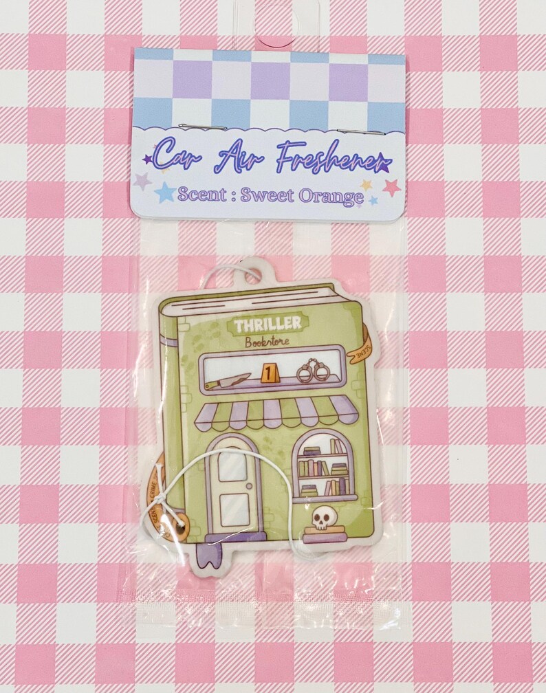 May include: Car air freshener in a clear plastic package. The air freshener is shaped like a green bookstore with the text "Thriller Bookstore" and "Scent: Sweet Orange". The background is a pink and white checkered pattern.
