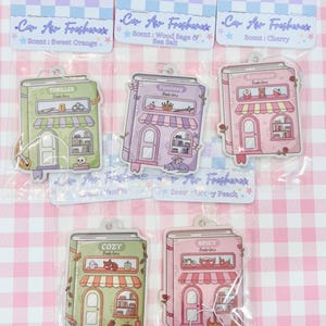 May include: Air fresheners in the shape of illustrated book storefronts. Each has a different theme: Thriller, Fantasy, Romance, Cozy, and Spicy. The packaging reads "Car Air Freshener" and lists scents like Sweet Orange and Cherry.