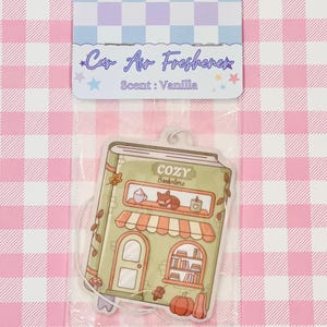 May include: Car air freshener in a clear plastic package. The air freshener is shaped like a book-shaped storefront with the text "COZY Bookstore". The scent is vanilla. The background is a pink and white checkered pattern.