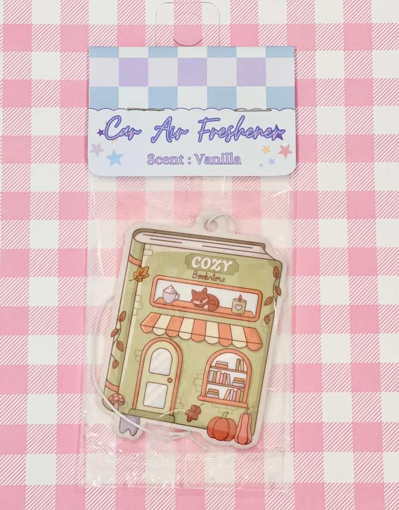 May include: Car air freshener in a clear plastic package. The air freshener is shaped like a book-shaped storefront with the text "COZY Bookstore". The scent is vanilla. The background is a pink and white checkered pattern.