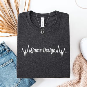 May include: Dark grey heather t-shirt featuring the text "Game Design" in a heartbeat graphic. The shirt is folded, alongside blue jeans, a necklace, and white trainers.