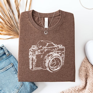 May include: A heather brown t-shirt with a white graphic of a vintage camera. The shirt is folded, with a silver necklace and a pair of blue jeans and white shoes nearby. The shirt has the words "Heather Brown" on it.