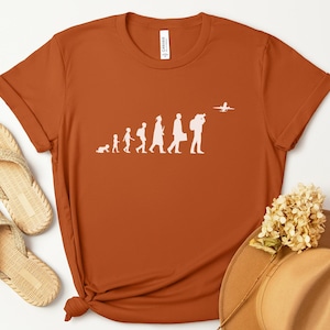 May include: Rust-colored t-shirt with a white graphic depicting the evolution of travel, from crawling to boarding a plane. The word "Autumn" is in a circular orange design. Accessories include woven sandals and a tan hat.