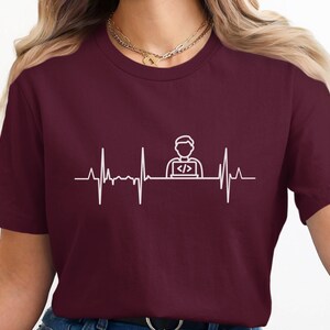 May include: Maroon t-shirt with a white graphic depicting a heartbeat line and a person with a laptop. The short-sleeved shirt is made of a soft material. The word 'Maroon' is in a circle.