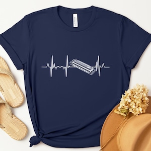 May include: Navy blue t-shirt featuring a white graphic of a harmonica integrated into a heartbeat line. The design suggests a musical theme. The shirt is made of a soft material and is suitable for casual wear.