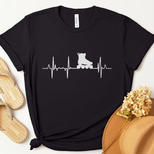 May include: Black t-shirt featuring a white roller skate design integrated into a heartbeat graphic. The shirt is paired with beige sandals and a brown hat with a floral accent, creating a casual, sporty aesthetic.