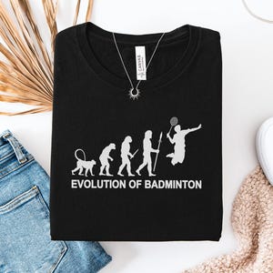 May include: A black t-shirt featuring a white graphic depicting the evolution of humans, ending with a person playing badminton. The text below the graphic reads "EVOLUTION OF BADMINTON" in white capital letters. The shirt has a crew neckline.