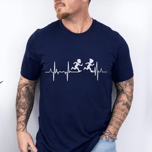 May include: Navy blue t-shirt featuring a white graphic of two running figures above a heartbeat line. The shirt is a solid color and has a crew neck. The word "Navy" is in a navy blue circle in the upper left corner.