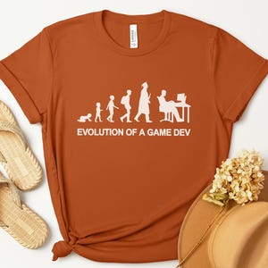 May include: An orange t-shirt with the text "EVOLUTION OF A GAME DEV" in white. The graphic shows a person's evolution from crawling to working at a computer. The shirt is next to a pair of sandals and a hat.