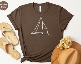 Peut inclure: T-shirt marron chiné avec un motif de voilier blanc. L'image comprend des sandales beiges, un chapeau beige et des fleurs séchées. Le t-shirt est fait d'une matière douce.
