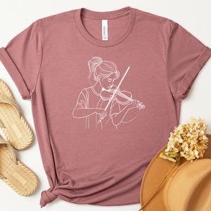 May include: Heather mauve t-shirt with a white outline of a person playing a violin. The shirt has rolled sleeves and a knot at the hem. Woven sandals and a wide-brimmed hat are also in the image.