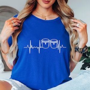 May include: Royal blue t-shirt featuring a white graphic of two bongos and a heartbeat line. The shirt has a crew neck and short sleeves. The text "True Royal" is in a blue circle in the upper left corner.