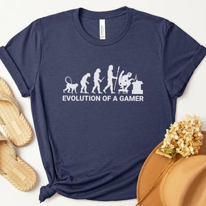 May include: Heather navy blue t-shirt with the white text "EVOLUTION OF A GAMER" and a graphic depicting the evolution of humans into gamers. The shirt is paired with beige sandals and a brown hat.