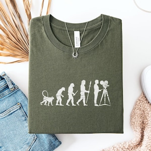 May include: Military green t-shirt with a white graphic showing the evolution of a filmmaker, from ape to person with a camera. The shirt is folded on a white surface, next to blue jeans and white trainers.