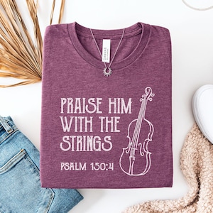 May include: Heather maroon t-shirt with the text "Praise Him With The Strings" and a violin illustration. Psalm 150:4 is printed below the text. A silver necklace and white shoes are also visible.
