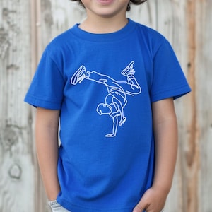 May include: A royal blue t-shirt featuring a white line drawing of a breakdancer in a handstand pose. The shirt is short-sleeved and has the words "True Royal" in a blue circle in the upper left corner. The background is a wooden wall.