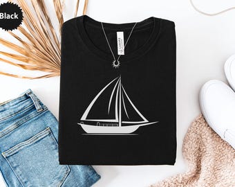 Peut inclure: T-shirt noir avec un motif de voilier blanc. Le t-shirt a un col rond et des manches courtes. Un collier argenté avec un pendentif soleil est visible. Le t-shirt est posé sur une surface blanche à côté d'un jean bleu et de baskets blanches.