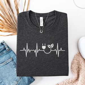 May include: Dark gray heather t-shirt with a white graphic of a heartbeat line, a plug, and a leaf. The shirt is folded and displayed with blue jeans, a necklace, and white shoes.