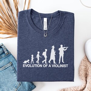 May include: Navy blue t-shirt featuring the white text "EVOLUTION OF A VIOLINIST". The graphic depicts the stages of a violinist's life. A silver necklace and a pair of blue jeans are also visible in the image.