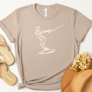 May include: A tan-coloured t-shirt with a white line drawing of a wakeboarder. The shirt is styled with woven sandals and a brown hat with a dried flower. The word "Tan" is in a tan circle in the upper left corner.