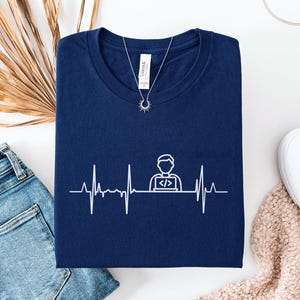 May include: Navy blue t-shirt with a white graphic of a heartbeat line and a programmer icon. The shirt is folded, with a silver necklace and a pair of white shoes visible. A pair of blue jeans and a decorative plant are also in the image.