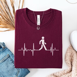 May include: Maroon t-shirt with a white graphic of a person walking within a heartbeat line. The shirt is paired with blue jeans, white shoes, and a necklace. A dried palm leaf is also in the image.