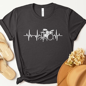 May include: Dark grey heather t-shirt with a white heartbeat graphic featuring a drum set. The shirt has a crew neck and short sleeves. The text "Dark Grey Heather" is in a circle in the upper left corner.