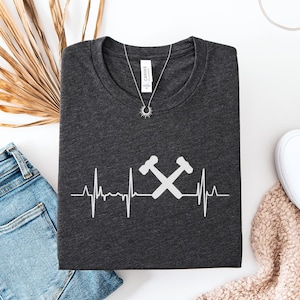 May include: Dark gray heather t-shirt with a white heartbeat graphic featuring crossed hammers. The shirt is folded and displayed with blue jeans, white shoes, and a necklace. The text "Dark Grey Heather" is in the upper left corner.
