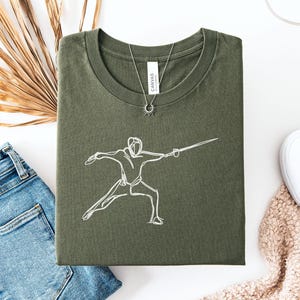 May include: Military green t-shirt with a white line drawing of a fencer in action. The shirt is folded and displayed with blue jeans, white sneakers, and a necklace. The word "Canvas" is visible on the tag.