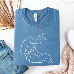 May include: Steel blue t-shirt with a white line drawing of two people in a martial arts move. The shirt is folded and paired with blue jeans, white sneakers, and a necklace. A dried palm leaf is also in the image.