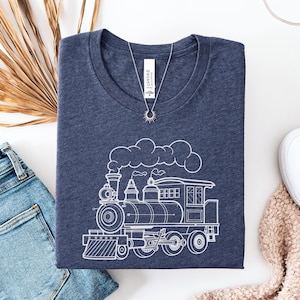 May include: Heather navy t-shirt with a white outline of a steam train. The shirt has a crew neck and short sleeves. A silver necklace with a sun pendant is on the shirt. The shirt is folded and placed on a white surface with blue jeans and white shoes.