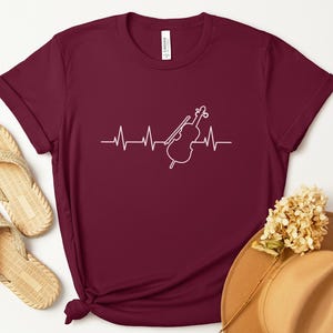 May include: Maroon t-shirt featuring a white heartbeat line design with a cello graphic. The shirt is a short-sleeved crew neck. The image also includes a pair of beige sandals and a tan hat.